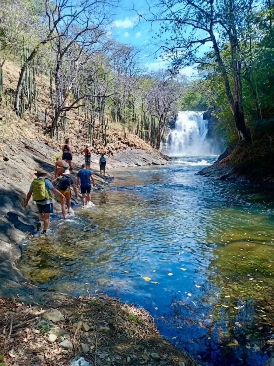 Hikers wading through a river toward a waterfall in the dry forest